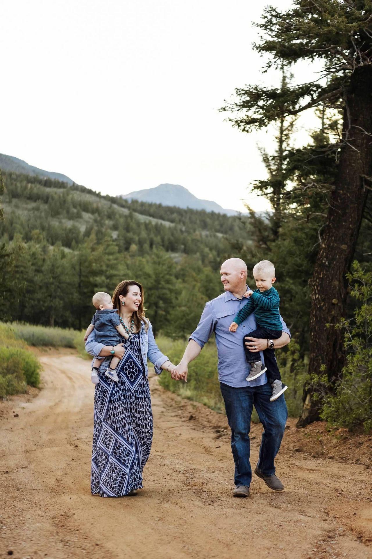 Family walking on a dirt path in a forested mountain area, carrying two young children.